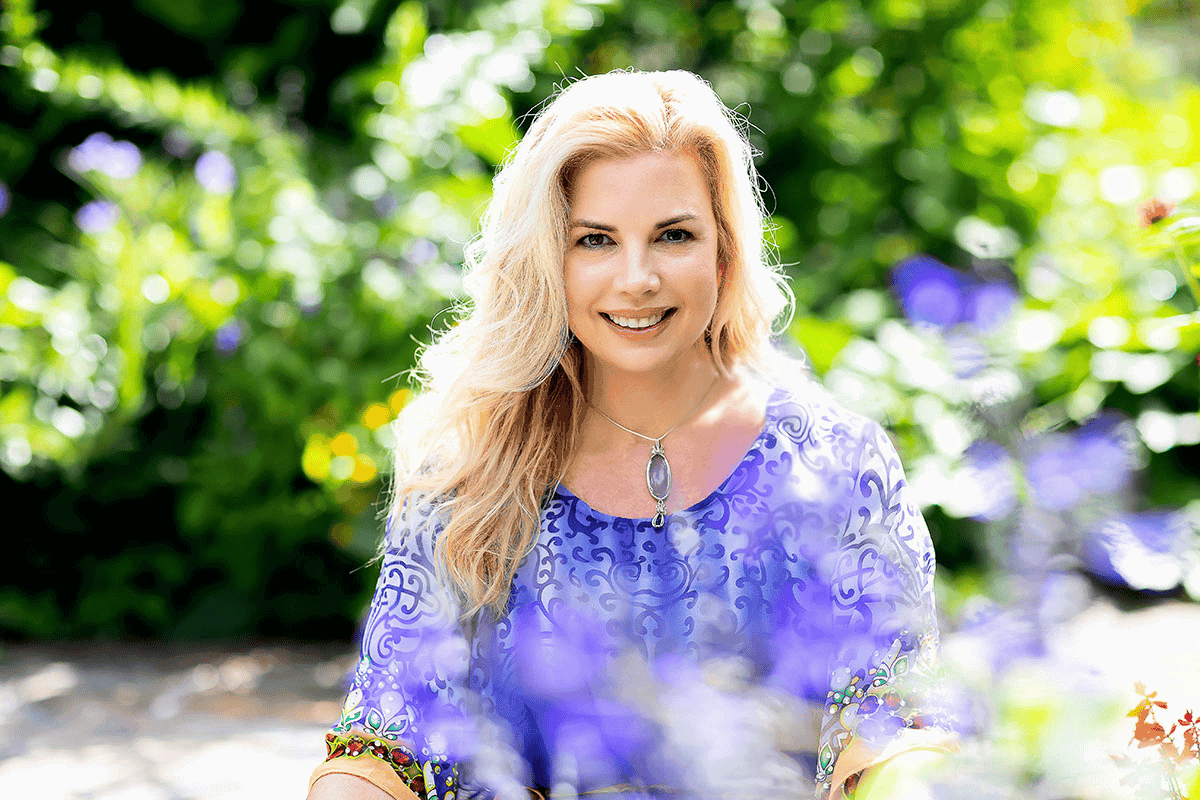 Headshot of Tracy, a confident woman with a warm smile. She has shoulder-length blonde hair and is sitting amongst vibrant flowers. Tracy is wearing a beautiful, stylish top that complements the colorful floral background, highlighting her friendly and approachable demeanor.